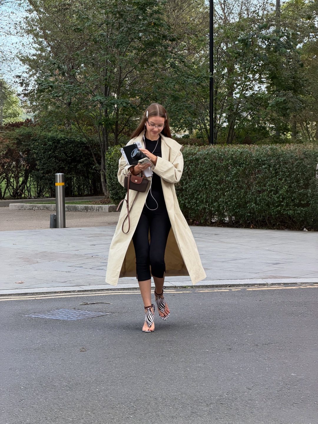 Klaudija in an ivory trench coat, walking with her phone and a camera box